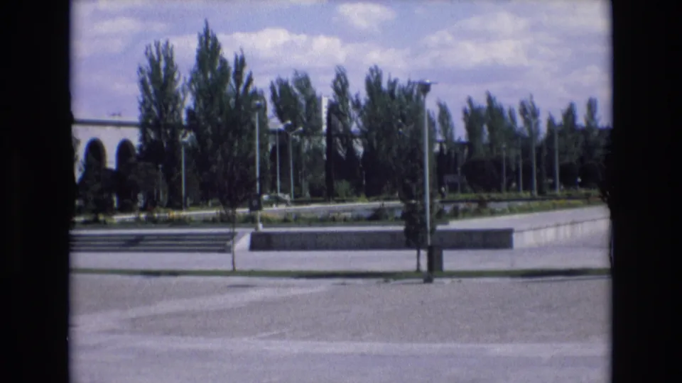 1972 Spain home-movie frame showing a broad paved plaza with tall trees, modernist arches at left, and a bright partly cloudy sky.