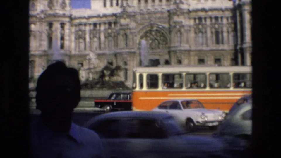 1972 Spain street scene with a silhouetted person in the foreground, an orange-and-cream tram, cars, and an ornate stone building behind them.