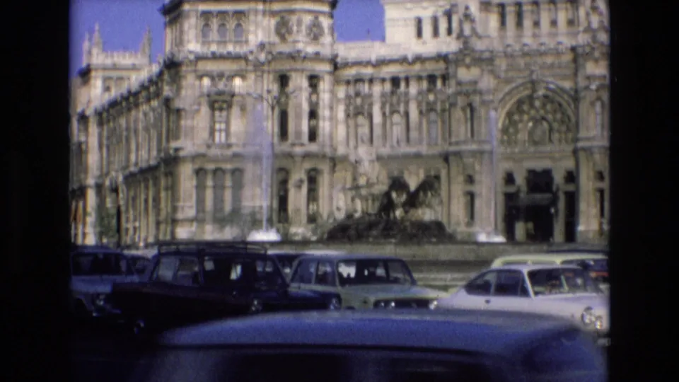 1972 Spain frame showing an ornate stone facade, a central fountain, and several classic cars passing or parked in the foreground.