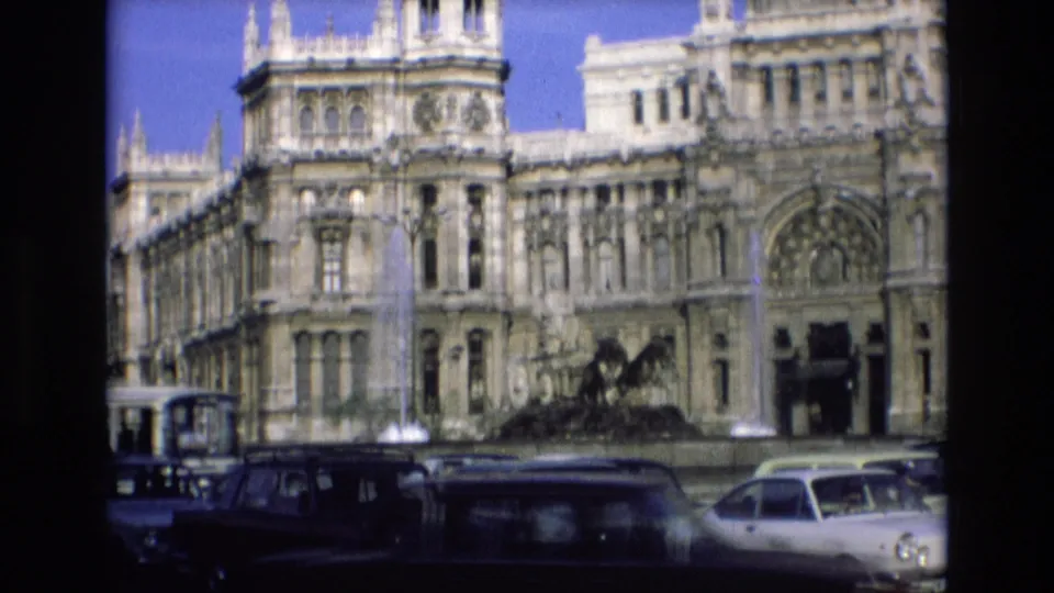 1972 Spain home-movie frame of a grand stone building and fountain seen behind classic cars, with part of a tram visible at the left edge.