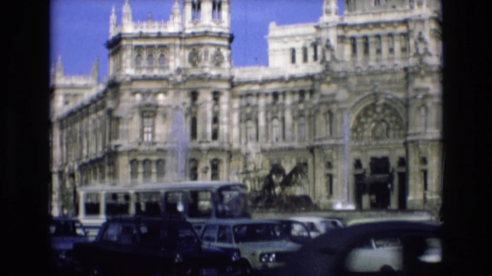 1972 Spain home-movie frame showing classic cars, a bus, and a fountain before a large ornate stone civic building under blue sky.