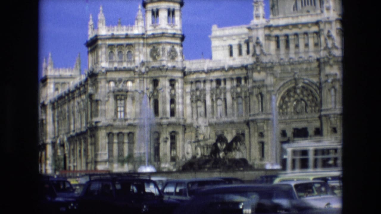 Classic cars and a bus move past a large historic-looking building and fountain.