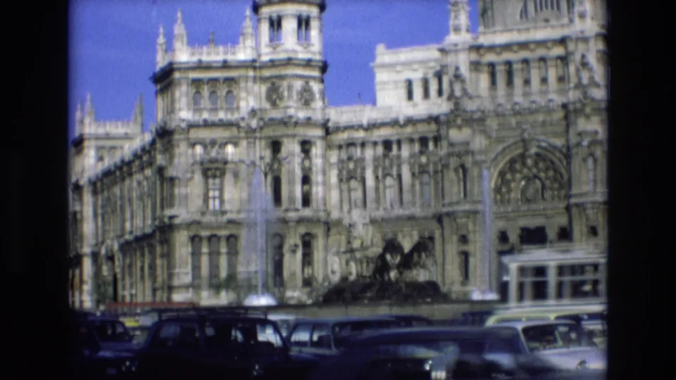 1972 Spain street view with mid-century vehicles passing a grand stone facade, fountain spray, and dark foreground frame edge from the film scan.