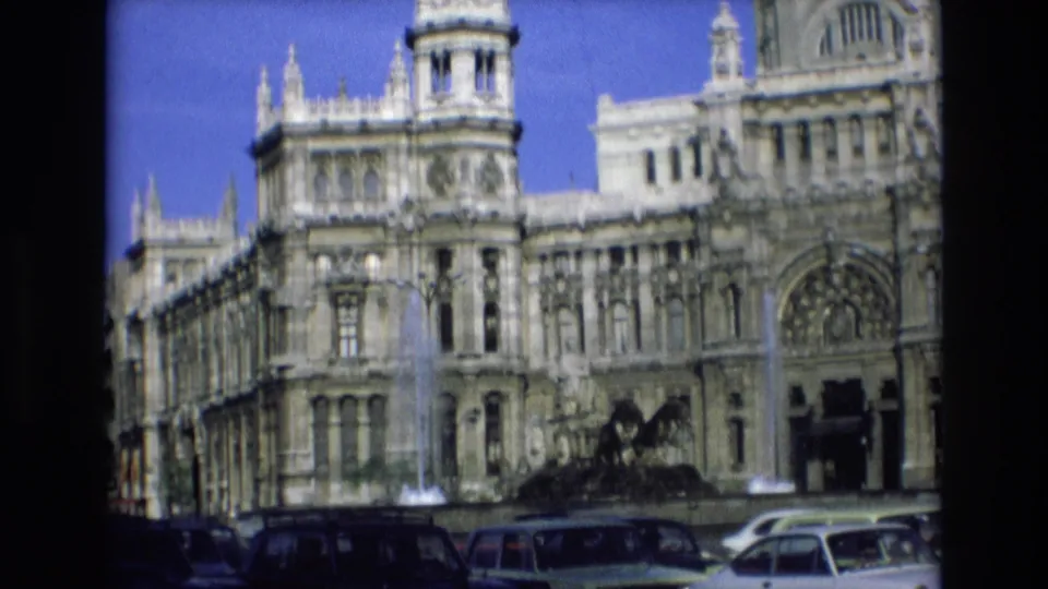 1972 Spain archival frame of an ornate stone building with arched windows, towers, a fountain, and cars along the street foreground.