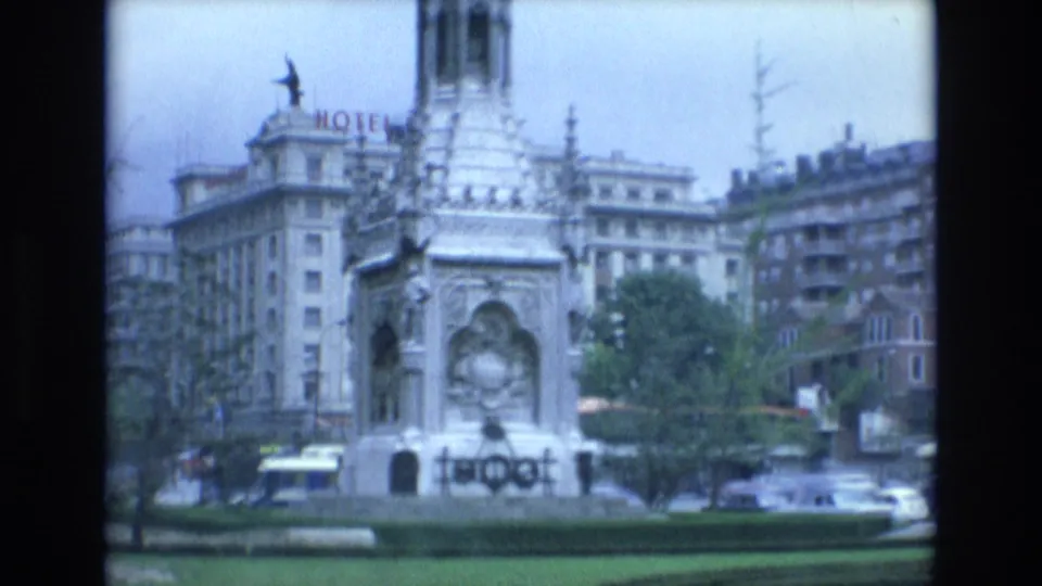 1972 Spain home-movie frame showing a public monument or fountain in a traffic circle, with surrounding city buildings, cars, greenery, and a hotel sign in the distance.