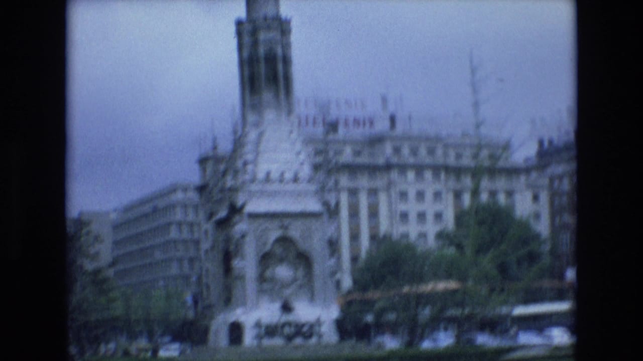 1972 Spain 8mm frame showing a tall stone monument beside a large building with visible hotel signage under an overcast sky.