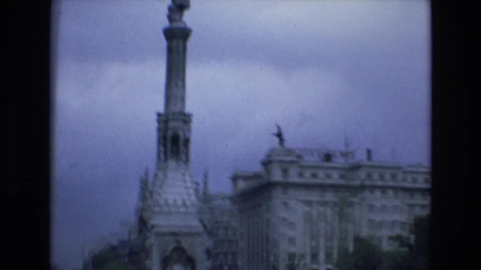 1972 Spain 8mm frame with the upper section of a stone column monument and a large multi-story building visible through motion blur.