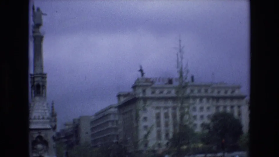 1972 Spain 8mm frame showing a moving-car view past the side of the monument, adjacent buildings, trees, and overhead wires.