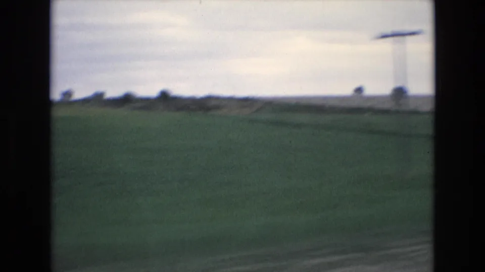 1972 Spain 8mm frame looking across green roadside fields with distant utility poles and wires beneath a pale cloudy sky.