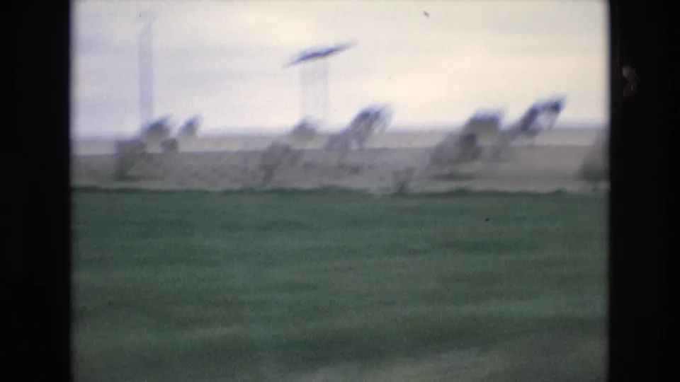 Soft 8mm view of rural Spain in 1972 with a green foreground, distant trees, a power line tower, and motion blur from travel.
