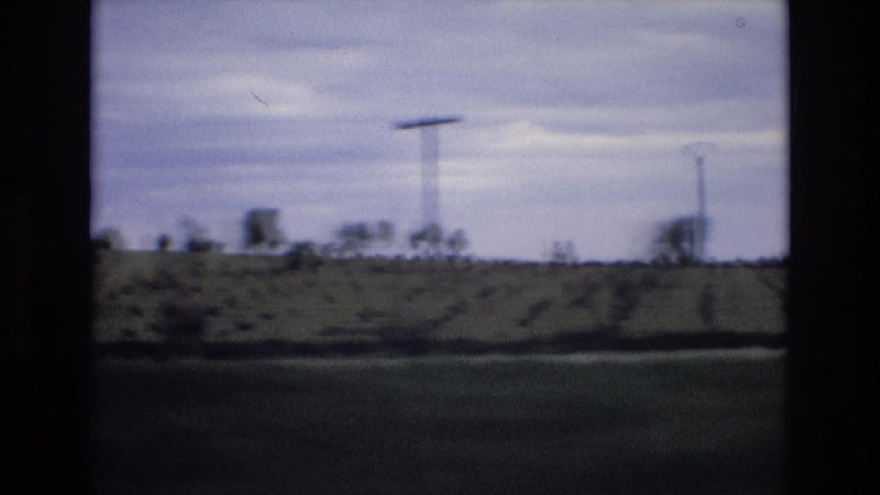 1972 Spain home-movie frame showing blurred rural fields, utility poles, and a low tree line from a moving vehicle under a cloudy sky.