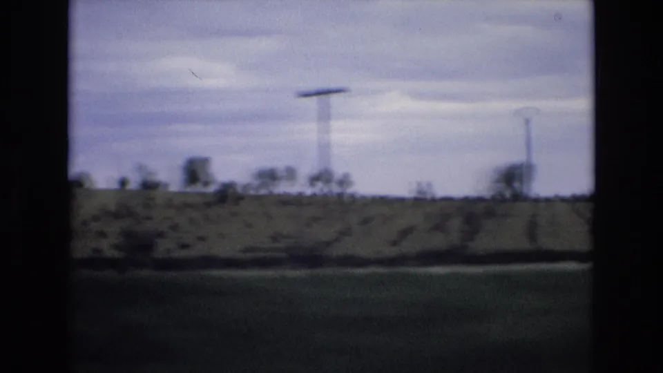 1972 Spain home-movie frame showing blurred rural fields, utility poles, and a low tree line from a moving vehicle under a cloudy sky.