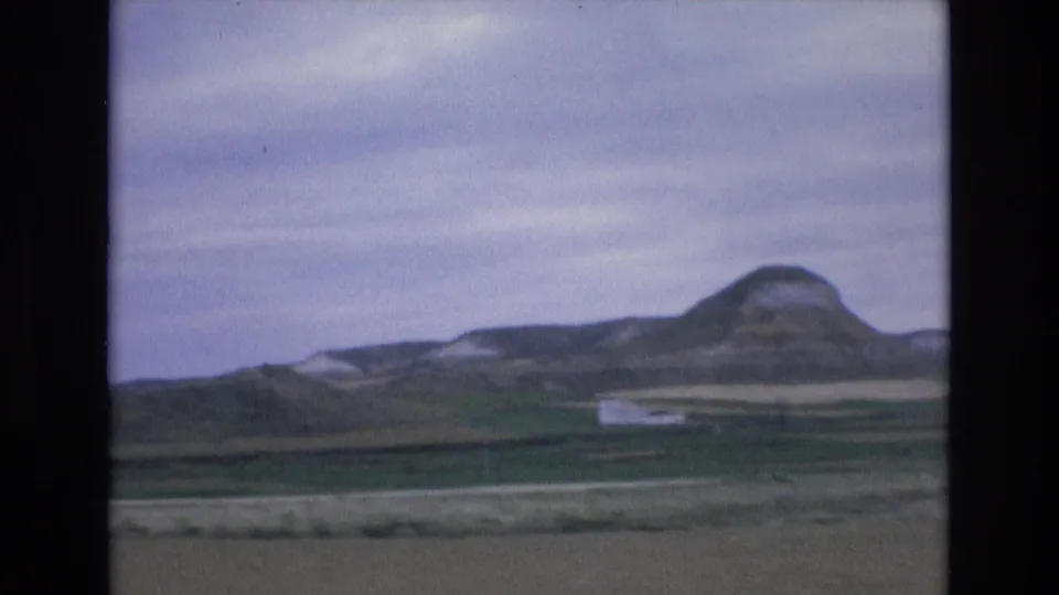 1972 Spain rural landscape frame with open fields, distant ridges, and a prominent rounded hill under an overcast sky.