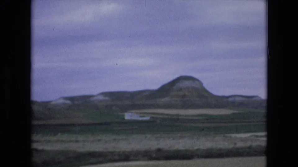 1972 Spain home-movie view of rolling farmland, a rounded hill, pale field bands, and a blurred roadside foreground from a moving vehicle.