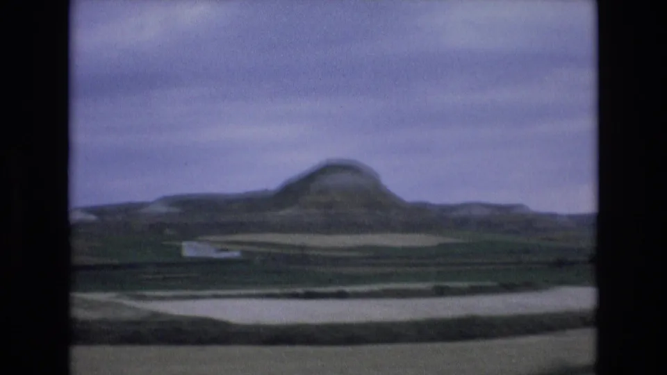 1972 Spain rural landscape frame showing a distant rounded hill, layered fields, and pale roadside ground beneath a muted overcast sky.