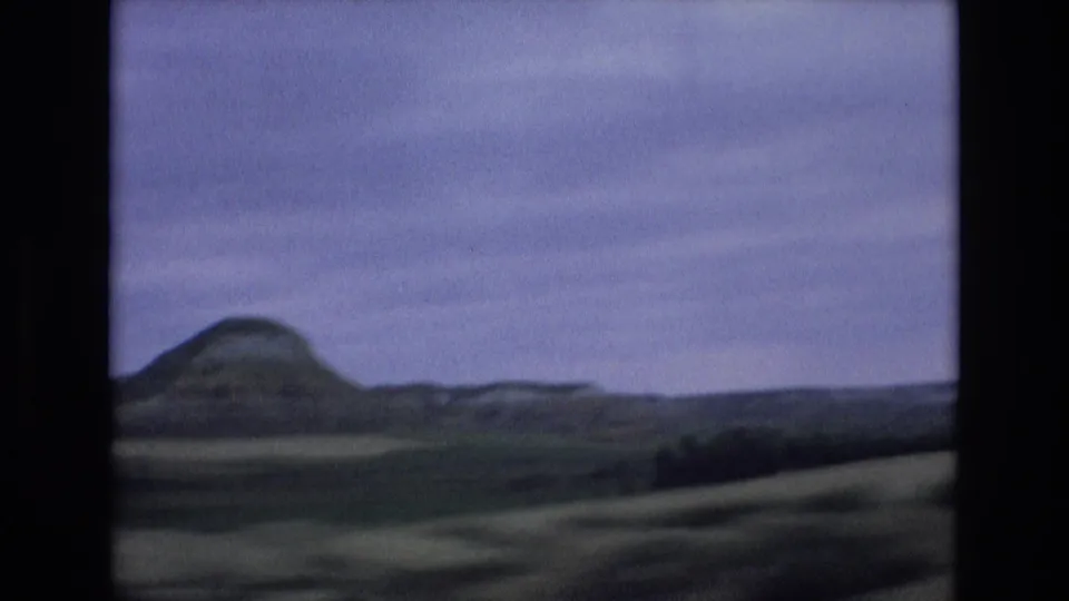 1972 Spain travel frame with a distant rounded mountain, rolling farmland, and motion-softened foreground from a moving viewpoint.