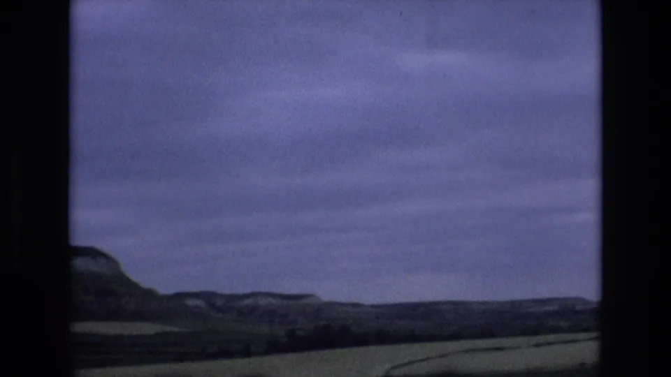 Soft 8mm frame from the Spain clip showing low hills and broad fields under a purple-blue overcast sky.