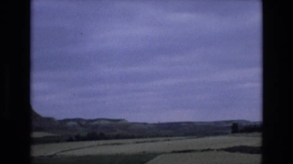 1972 home-movie frame from Spain showing a wide rural valley, pale fields, and distant low hills under an overcast sky.