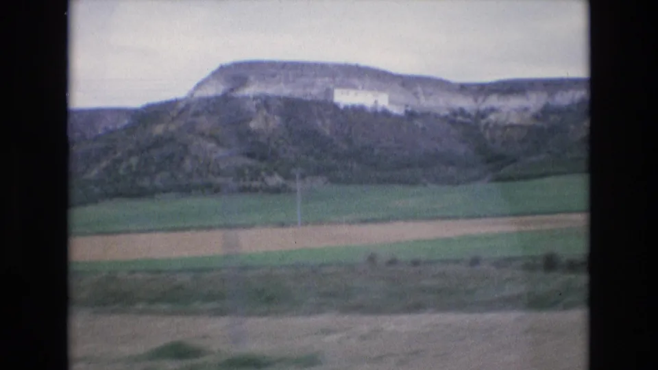1972 Spain 8mm travel frame with green fields in the foreground, rocky plateau slopes, and a white hillside building.