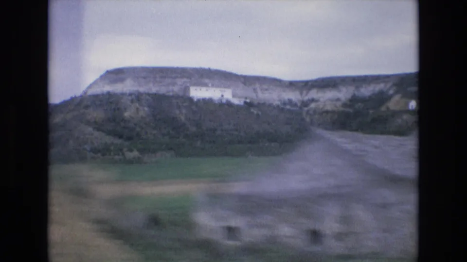 1972 Spain 8mm frame from a moving vehicle, with motion blur across fields, road surface, rocky hillside, and a white building.