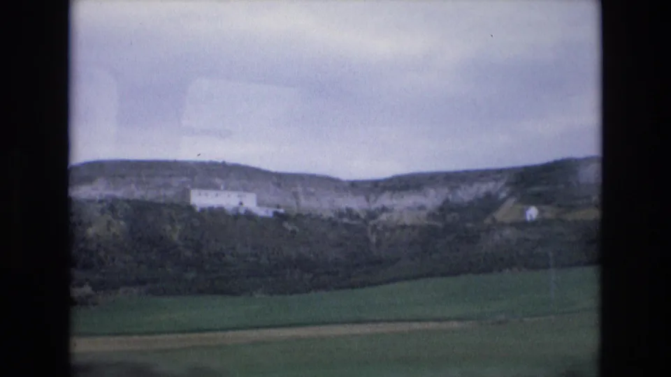 1972 Spain 8mm travel frame showing a green field, dark vegetation, rocky plateau, and white hillside building beneath a muted cloudy sky.