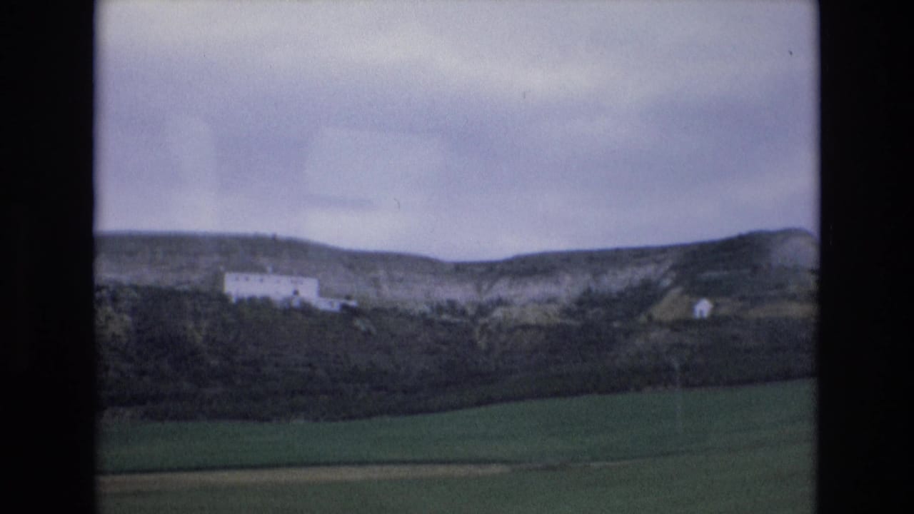 1972 Spain 8mm frame showing green fields, dark hedgerow, a distant white hillside building, and an overcast sky.