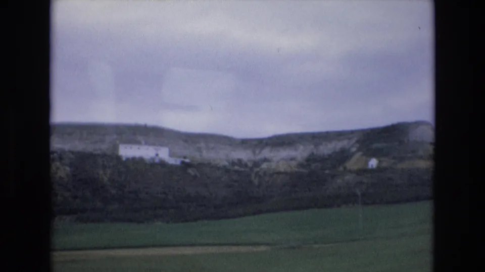 1972 Spain 8mm frame showing green fields, dark hedgerow, a distant white hillside building, and an overcast sky.