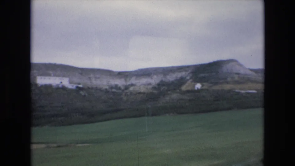1972 Spain 8mm frame with open green foreground, hillside scrub, small white buildings, and a broad cloudy sky.