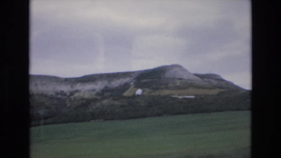 1972 Spain 8mm frame showing a rocky rural hill, dark scrub line, green fields, and a small white building on the slope.