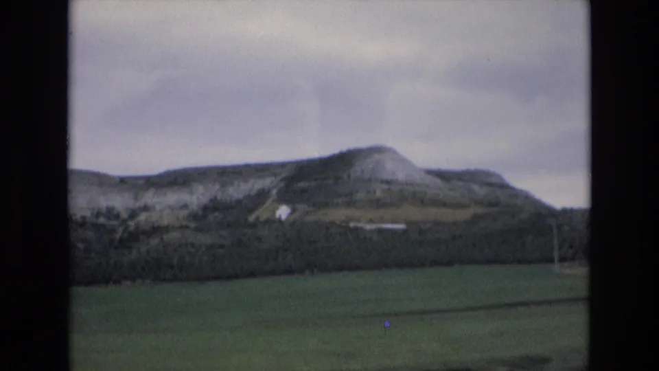 1972 Spain 8mm frame showing open green countryside, a rocky hill with a pale structure, dark vegetation, and muted cloudy light.