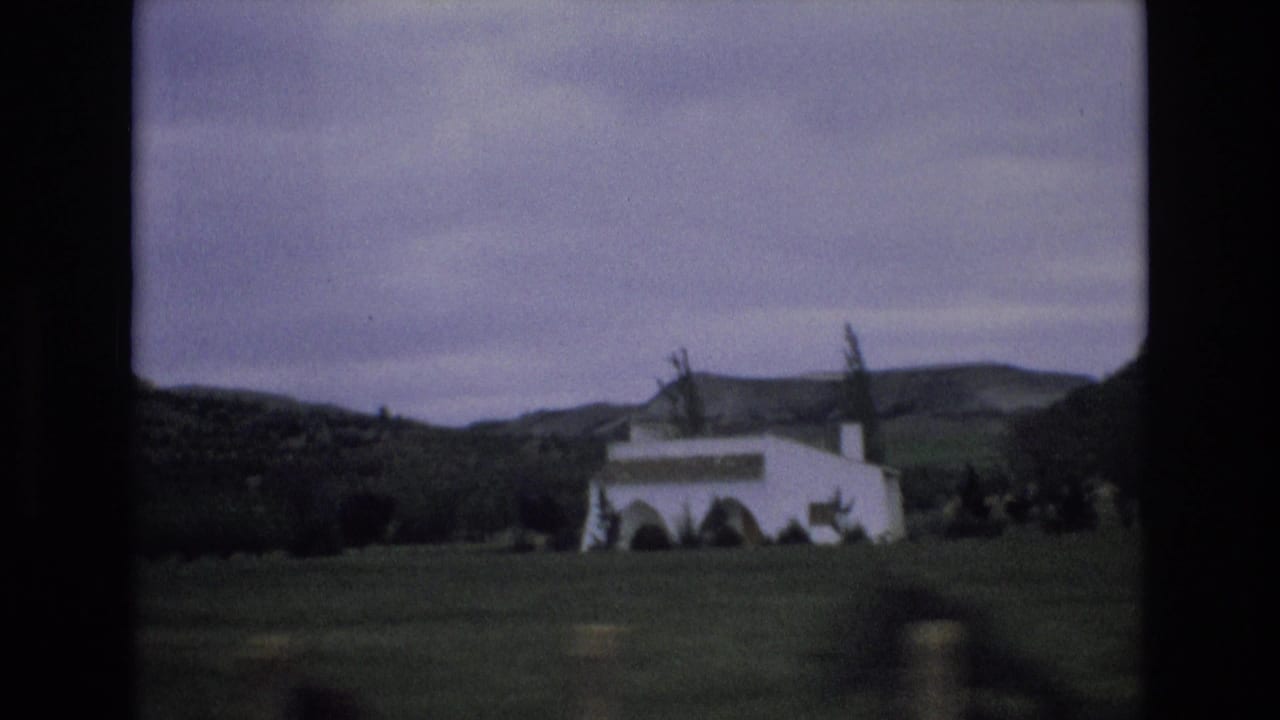 1972 Spain 8mm frame showing a white rural building in a valley, with trees, low hills, and a cloudy sky seen from a moving viewpoint.