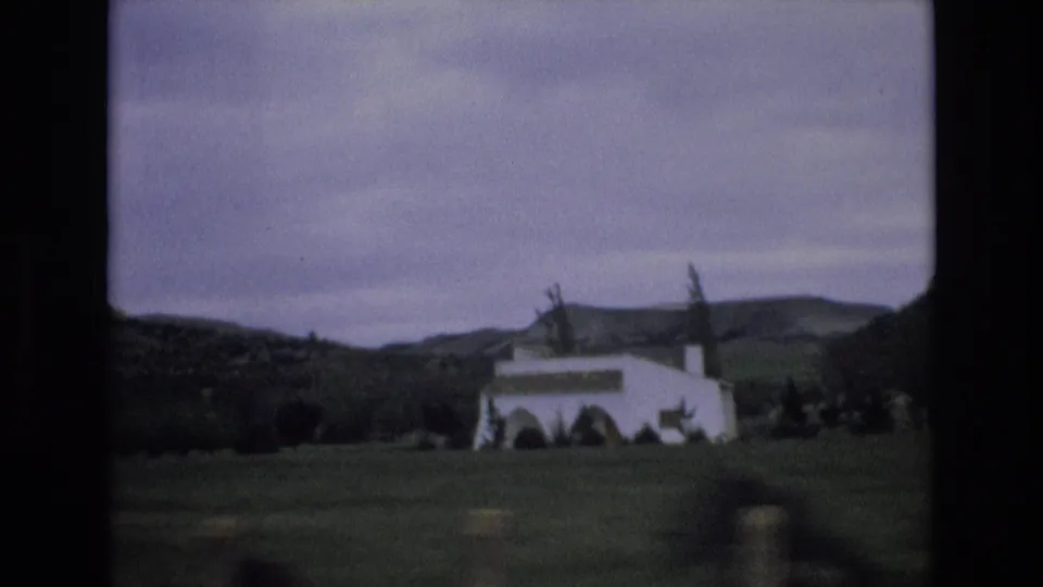 1972 Spain 8mm frame showing a white rural building in a valley, with trees, low hills, and a cloudy sky seen from a moving viewpoint.