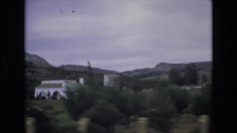 Moving 8mm view in Spain, 1972, with a white building, trees, foreground posts, and soft hills under a grey sky.