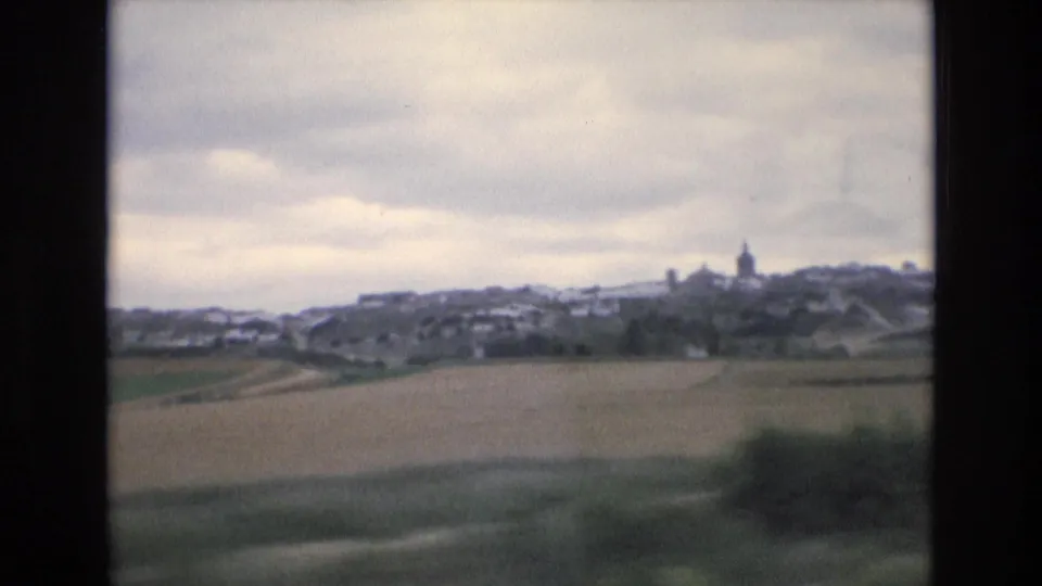 1972 Spain 8mm frame of a distant village across open fields, with clustered buildings and a church steeple beneath an overcast sky.