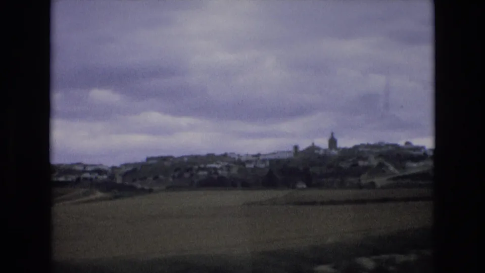 Wide 8mm travel view in Spain, 1972, showing fields, a distant village, a church steeple, and dark clouds over low hills.