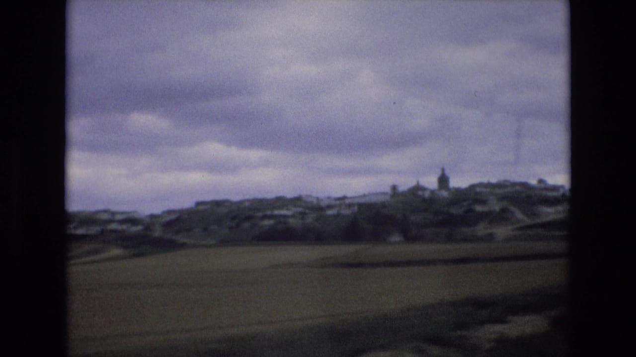 1972 Spain 8mm frame showing a green field in the foreground and a small hillside village with a steeple beneath overcast clouds.