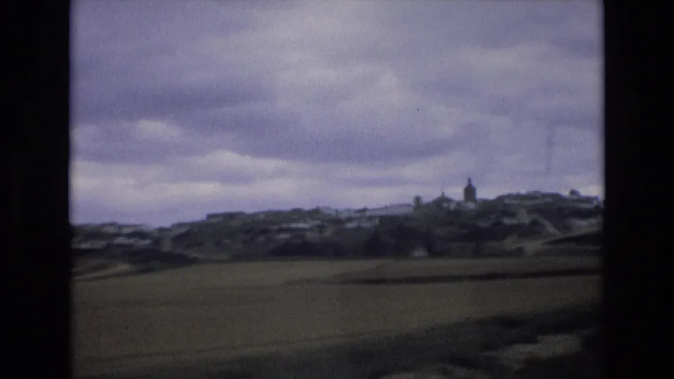 1972 Spain 8mm frame showing a green field in the foreground and a small hillside village with a steeple beneath overcast clouds.