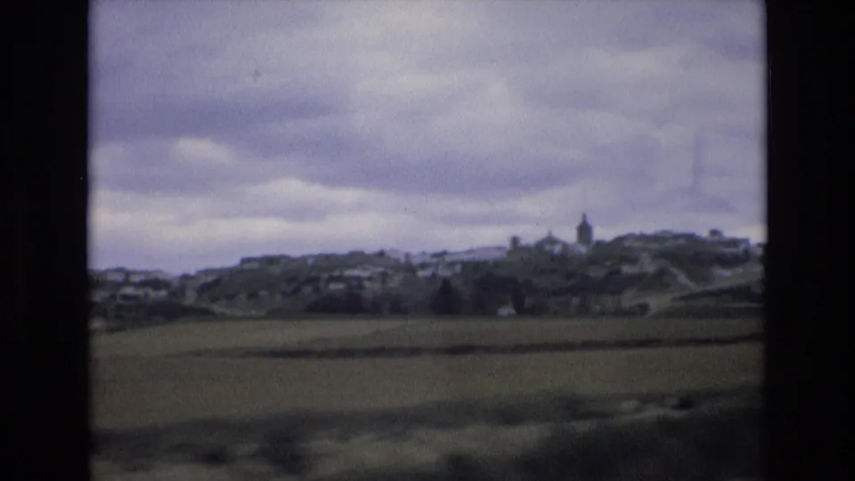 1972 Spain 8mm frame with open farmland leading toward a distant village and a visible steeple under layered gray clouds.