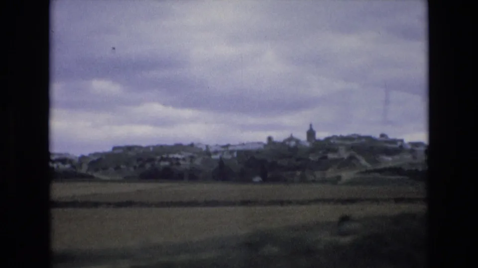1972 Spain 8mm frame looking across a green field toward a small hillside settlement with pale buildings and a steeple.