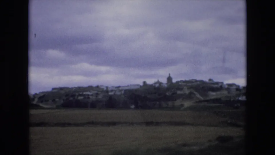 1972 Spain 8mm frame showing a cloudy rural landscape with open fields, a hillside village, and a prominent steeple in the distance.