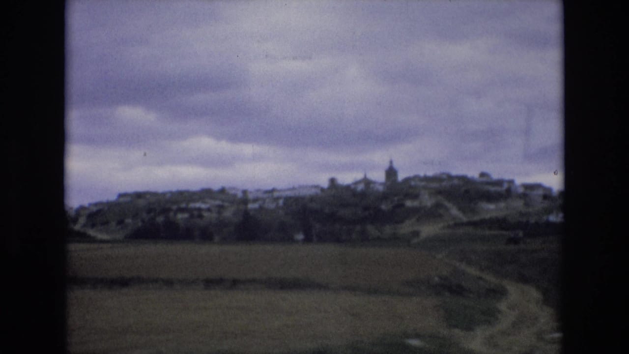 1972 Spain 8mm frame showing a distant hilltop village across open fields, with a dirt track and heavy cloud cover.