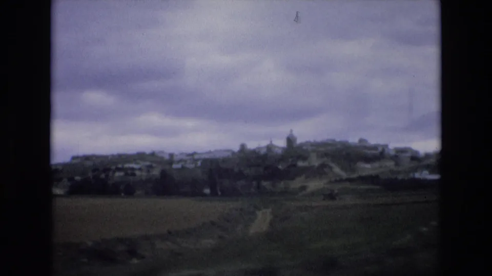 1972 Spain 8mm frame with a soft distant view of a hilltop settlement, farmland, and a winding pale track beneath cloudy sky.