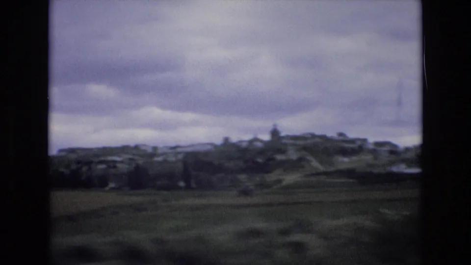 1972 Spain 8mm frame showing a blurred hilltop village above dark foreground fields, with layered clouds and no visible people.