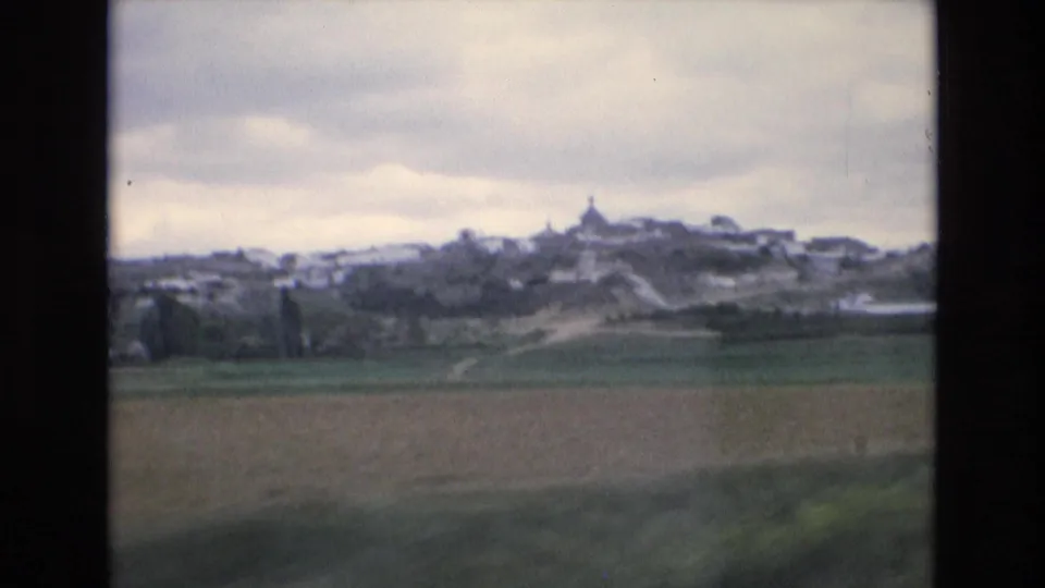 1972 Spain 8mm frame showing a hilltop village across green and brown fields, with a pale dirt path leading toward the settlement.