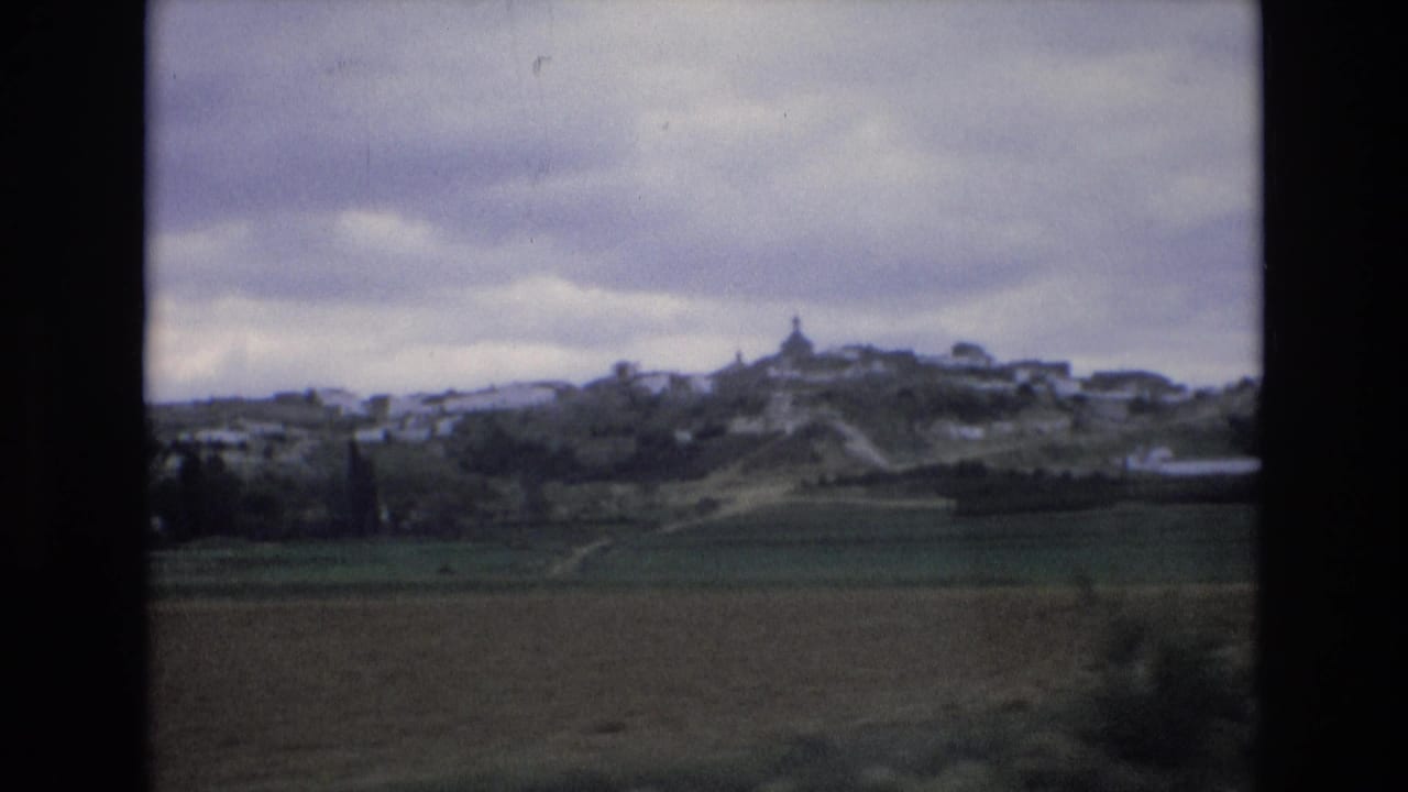 1972 Spain 8mm frame showing green and brown countryside with a distant white hill town and a small tower or steeple under heavy clouds.