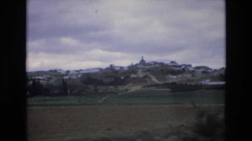 1972 Spain 8mm frame showing green and brown countryside with a distant white hill town and a small tower or steeple under heavy clouds.