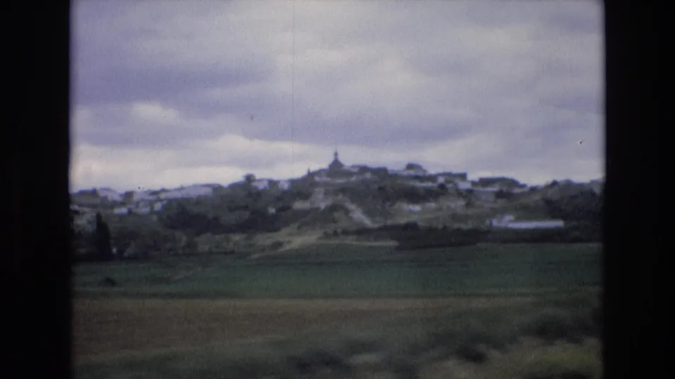 1972 Spain 8mm frame with a distant hillside settlement, open green fields, cloudy sky, and dark vertical framing from the moving train view.