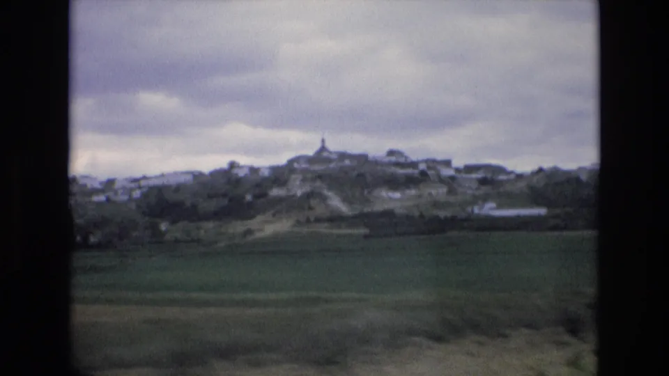 1972 Spain 8mm frame showing a wide rural foreground, distant white buildings clustered on a hill, and a small central tower beneath a gray sky.