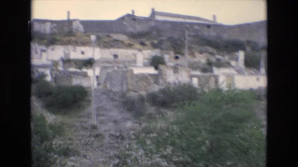 1972 Spain 8mm frame showing a closer passing view of white hillside buildings, low vegetation, and a long wall-like hilltop structure under pale sky.