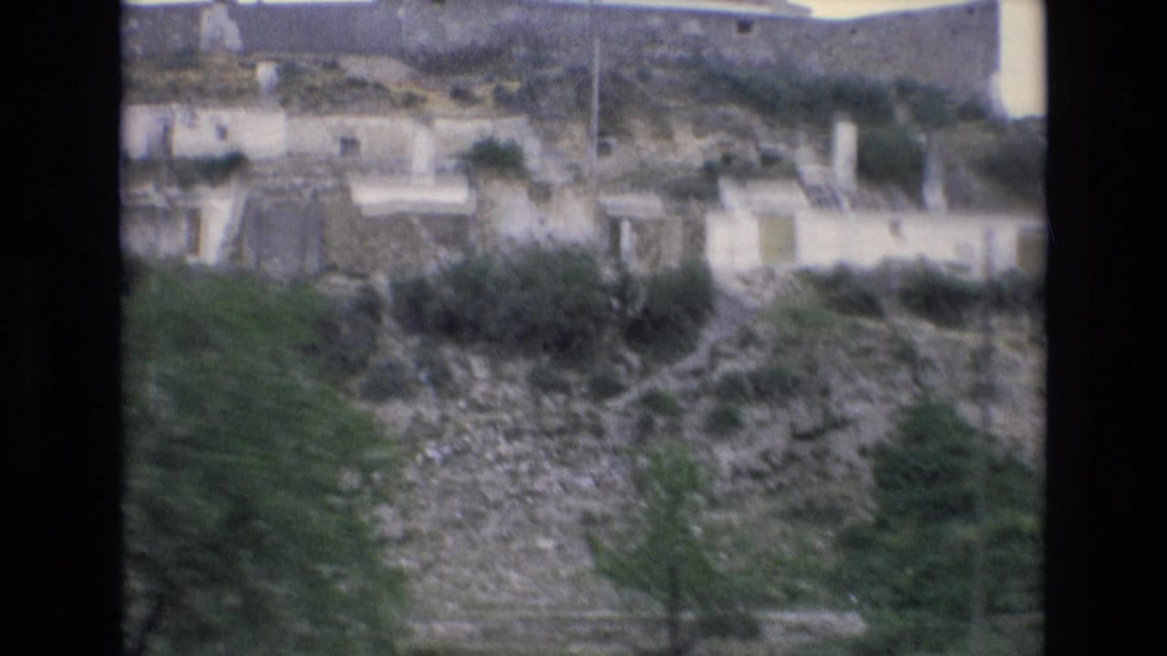 A 1972 Spain 8mm frame shows a distant rural hillside village with pale buildings, stonework, shrubs, trees, and rocky terrain under muted light.